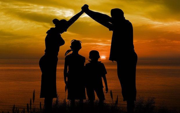 Save-A-Little_Life-Virtual -Classes-gerd-altmann-on-pixabay Shows an image of a family on the beach at sunset. A mother and father are holding their hands together creating an umbrella of protection above the heads of their children.