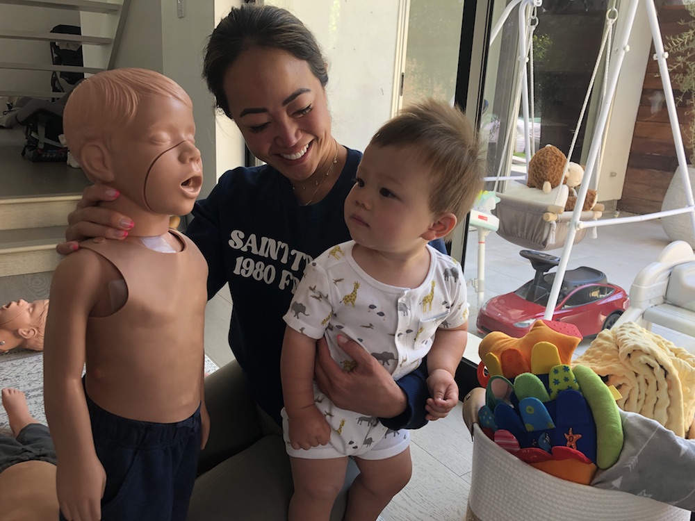 save-a-little-life-pediatric-cpr-and-family-safety Shows a kneeling woman who is holding her baby in one arm and a CPR mannequin in the other