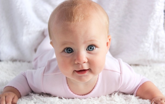 Shows a baby girl with bright blue eyes in a pink onesie laying on her stomach on top of a white shag rug. She is holding her head up and smiling while looking directly at the camera.