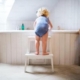 Shows a toddler on a small step stool climbing into a bathtub